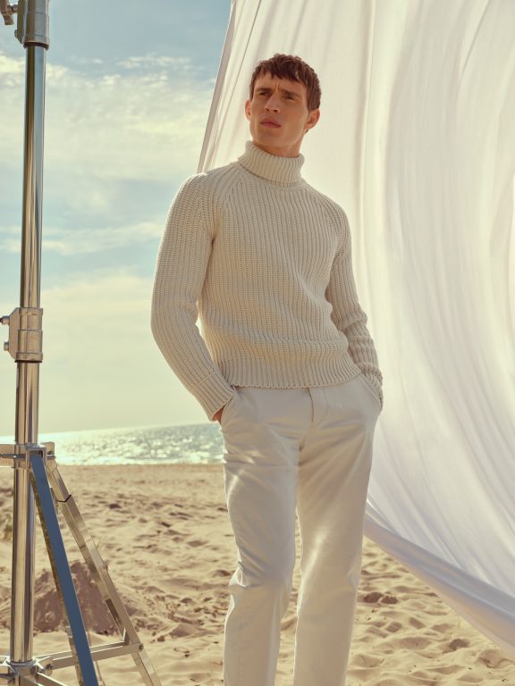 Mann in weißem Outfit steht vor Sonnensegel am Strand.