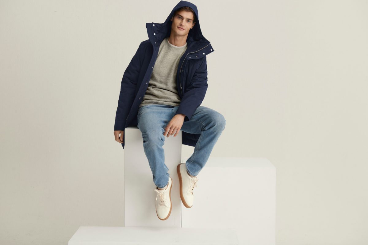 Male Model sitzt auf weißem Block in Parka und Jeans aus Naturfaser