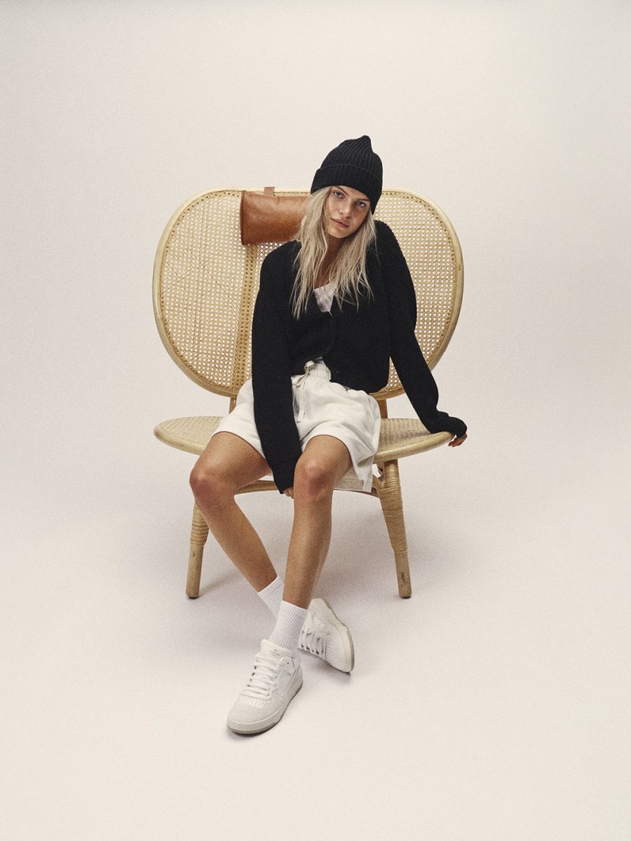 Female Model sitzt in Shorts, Strickjacke und Wollmütze auf Korbsessel.